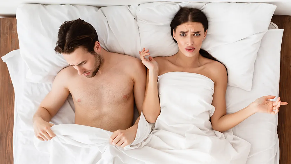 A man and a woman sit in bed under a white blanket; the man looks down, possibly troubled by low testosterone, while the woman looks up with a concerned expression.