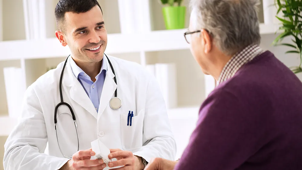 A doctor wearing a white coat and stethoscope smiles while discussing heart disease with an older man in a purple sweater in an office setting.