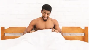 Shirtless man sitting up in bed, smiling and looking down at something under the white blanket, appreciating the surprising benefits of waking up hard.