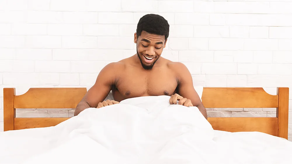 Shirtless man sitting up in bed, smiling and looking down at something under the white blanket, appreciating the surprising benefits of waking up hard.