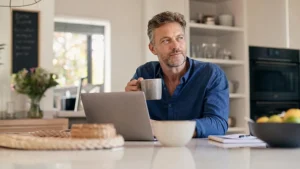 Man sitting at a kitchen counter with a laptop, holding a mug and looking to the side thoughtfully, perhaps researching Premature Ejaculation Treatments. A notebook, fruit bowl, and other kitchen items are visible.