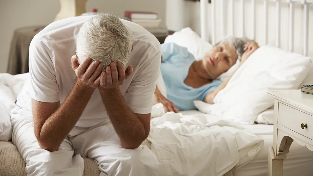 An older man sits on the edge of a bed with his head in his hands, possibly troubled by erectile dysfunction, while an older woman lies in bed behind him, looking concerned.