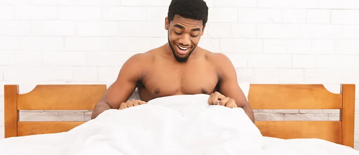 Shirtless man sitting up in bed, smiling and looking down at something under the white blanket, appreciating the surprising benefits of waking up hard.