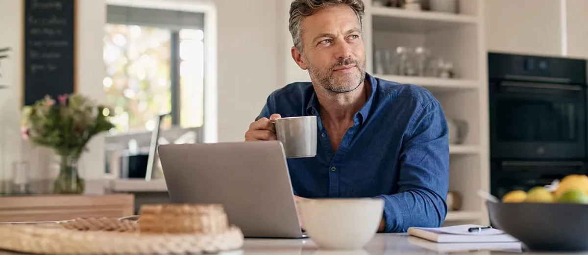 Man sitting at a kitchen counter with a laptop, holding a mug and looking to the side thoughtfully, perhaps researching Premature Ejaculation Treatments. A notebook, fruit bowl, and other kitchen items are visible.