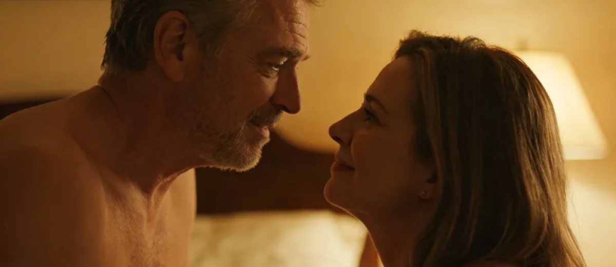 A shirtless middle-aged man and a woman sit close together on a bed, smiling and looking into each other's eyes in a warmly lit room, capturing the comfort and connection that can come from Sexual Performance Anxiety Treatment.